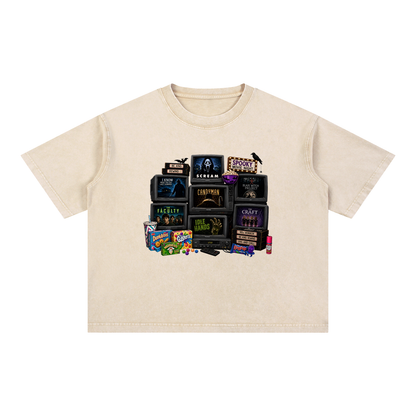A cream-colored short-sleeve t-shirt with a graphic print of vintage horror movie VHS tapes and candy, displayed flat against a solid white background.