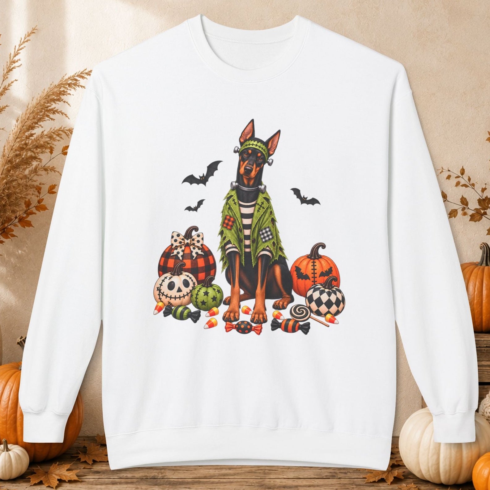 A white crewneck sweatshirt featuring a colorful graphic of a Doberman dog dressed in a green witch hat and cloak, surrounded by pumpkins, a skull, bats, and candy. The sweatshirt is displayed on a rustic wooden table surrounded by autumn decorations including pumpkins, dried grass, and a cozy knit blanket.
