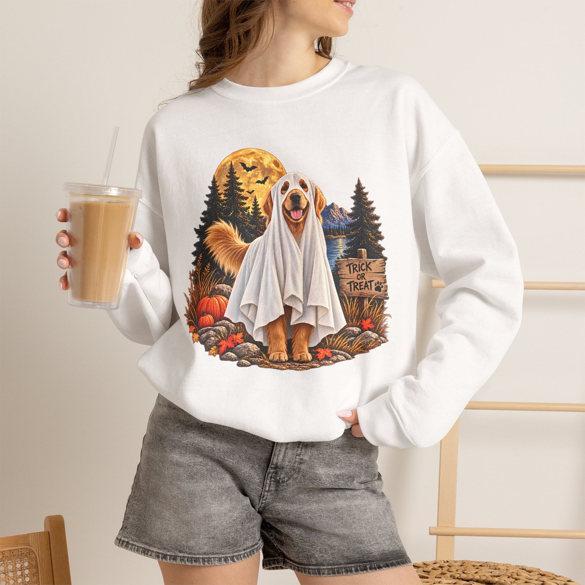 A woman wearing a white crewneck sweatshirt with a large graphic of a golden retriever dressed in a ghost costume surrounded by pumpkins, a full moon, and a sign that reads 'TRICK OR TREAT'. She is holding an iced coffee and wearing gray denim shorts.
