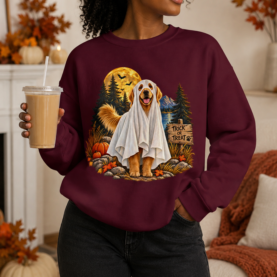 A woman wearing a burgundy sweatshirt with a graphic of a golden retriever dressed as a ghost in a spooky autumn scene, holding an iced coffee in a cozy living room setting.