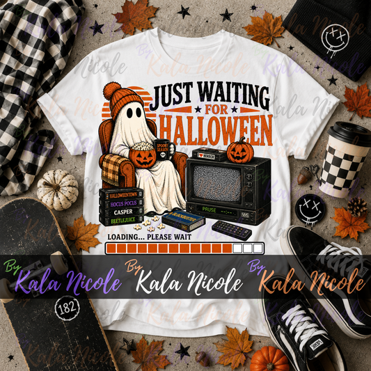 A white graphic T-shirt laid flat with Halloween-themed props around it, including a plaid scarf, skateboard, cup, sneakers, pumpkins, and autumn leaves. The shirt graphic shows a ghost in a pumpkin hat sitting in a plaid armchair next to a vintage TV, with text reading 'JUST WAITING FOR HALLOWEEN' and a loading bar.