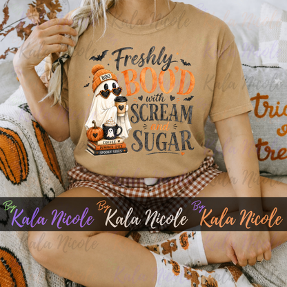 A woman wearing a tan graphic t-shirt with a Halloween ghost design and the text 'Freshly BOOED with SCREAM and SUGAR'. She is sitting on a cozy knit blanket with a gingham plaid skirt and patterned socks.